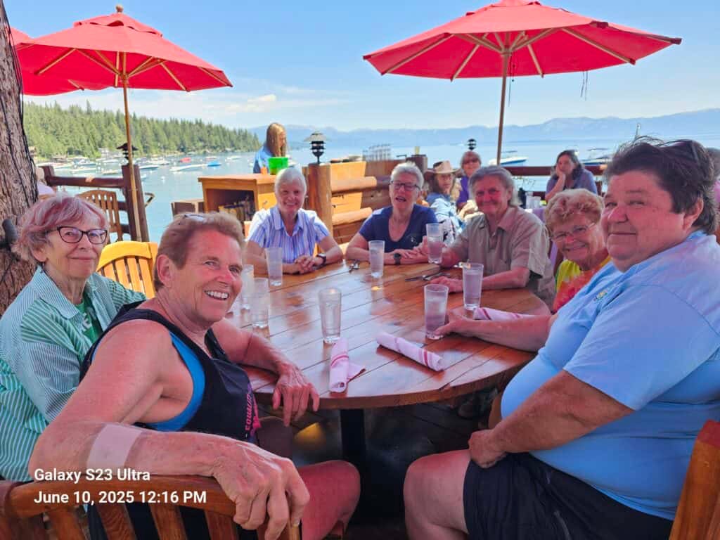 A group of smiling seniors enjoying a meal together at an outdoor lakeside restaurant with red umbrellas, with a scenic view of boats and mountains in the background, perfect for outdoor dining and social gatherings.